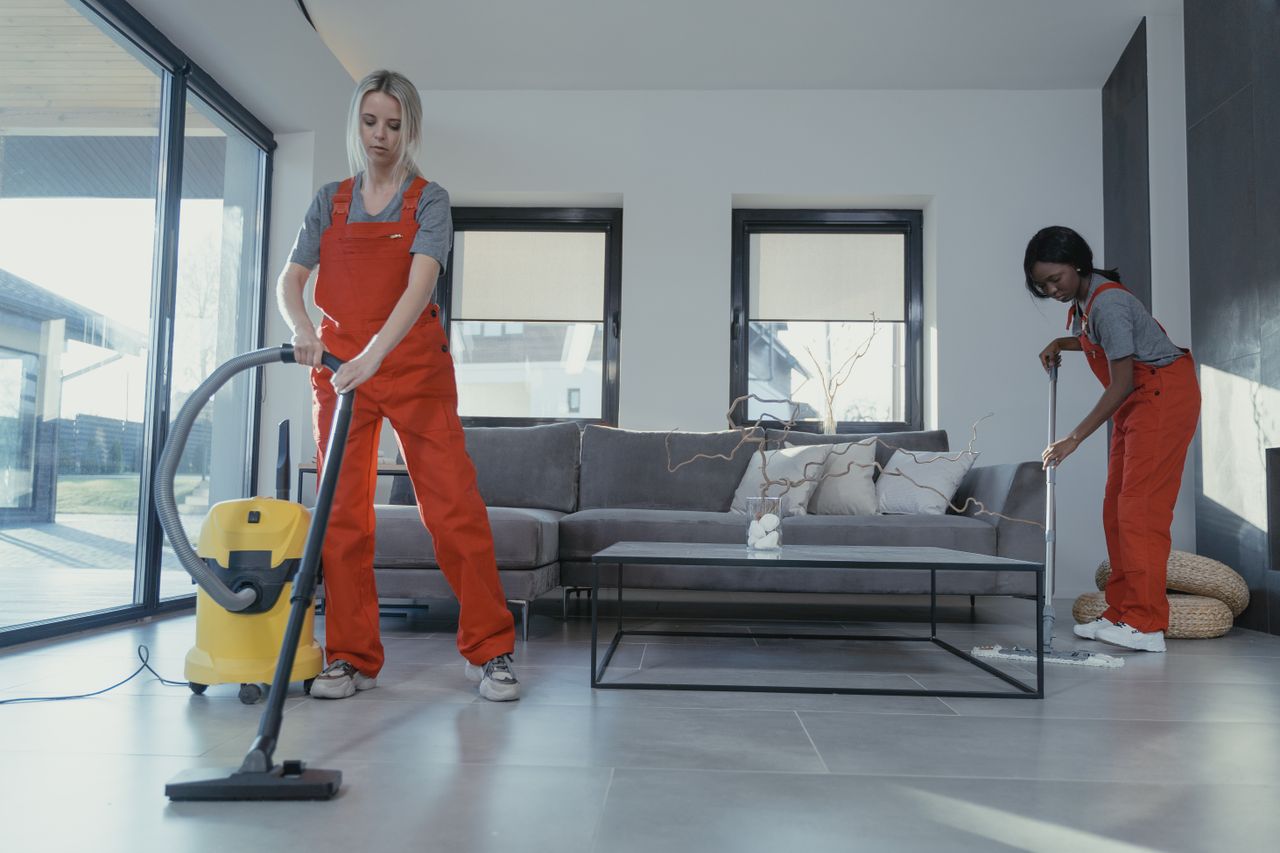 brand-02 Two women wearing red overalls are vacuuming and sweeping a modern living room.